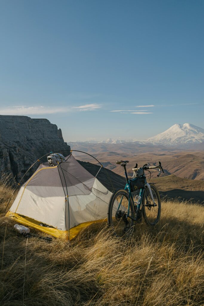 高原の草原に張られたテントと、そばに立つ旅装備の自転車。遠景には雪山がそびえる