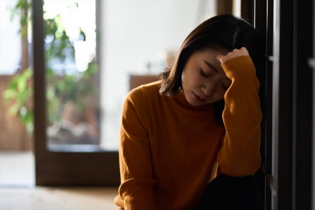 室内で床に座り、手で頭を支えながら目を閉じている女性。疲れた様子で、背後には自然光と観葉植物がある。