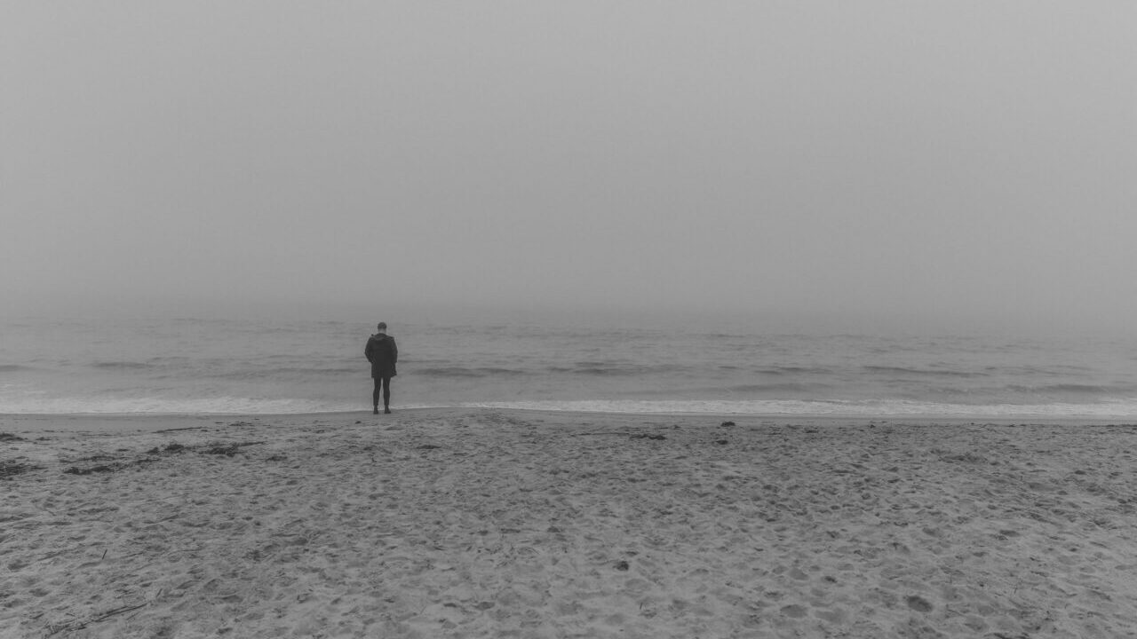 霧に包まれた海辺にひとり立つ人物。灰色の空と波が広がる静寂な風景。