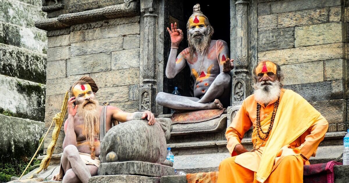 インドまたはネパールの石造りの寺院前でポーズをとる3人のサドゥ（ヒンドゥー教の修行者）。それぞれ顔や体に宗教的なペイントを施し、伝統的な装束を身にまとっている