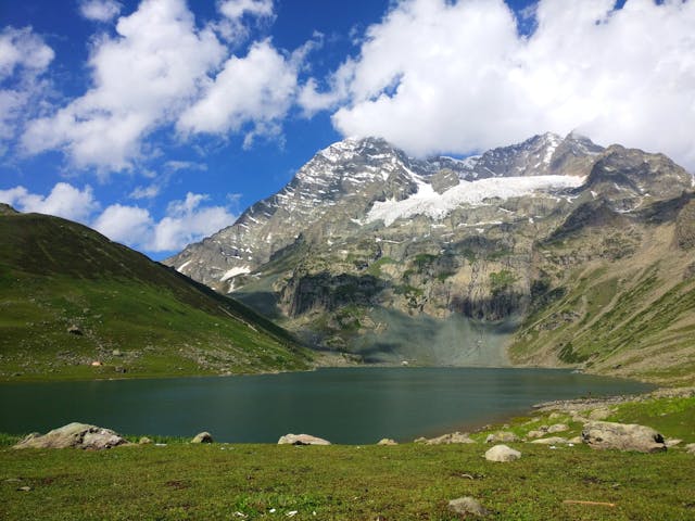 カシミールの山岳地帯に広がる氷河湖。背後には雪をまとった岩山がそびえ、手前には緑の草原が広がっている。