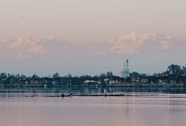 夕暮れのダル湖越しに見えるハズラトバル・モスクと、その背後に広がるヒマラヤの雪山。淡い空に浮かぶ静かな風景。